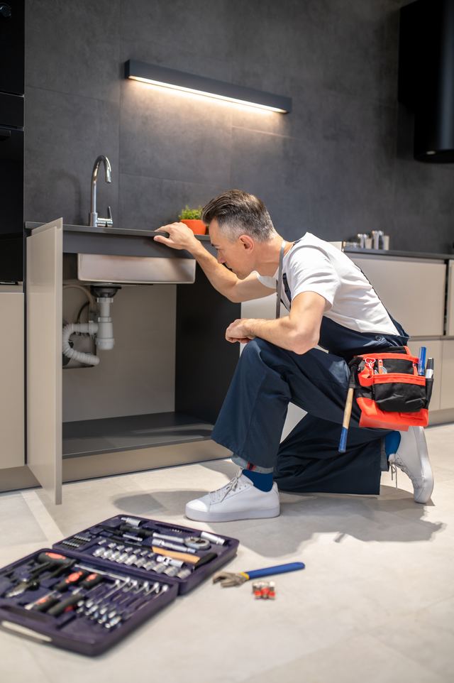 Fontanero arrodillado revisando las tuberías bajo un lavabo con su caja de herramientas